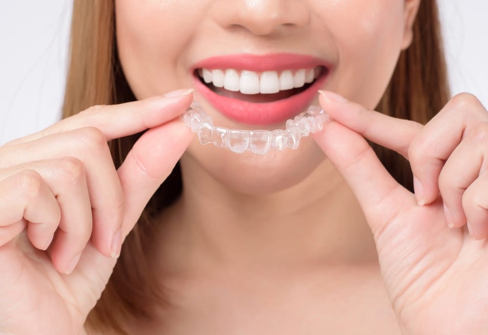 Smiling person holding a clear Invisalign aligner in front of their teeth, demonstrating removable clear aligners used to straighten teeth discreetly.