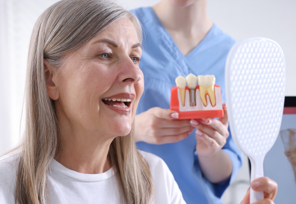 Smiling senior woman examining her smile in a mirror while a dental professional holds a dental implant model at Total Dental Arts in Centreville, VA.